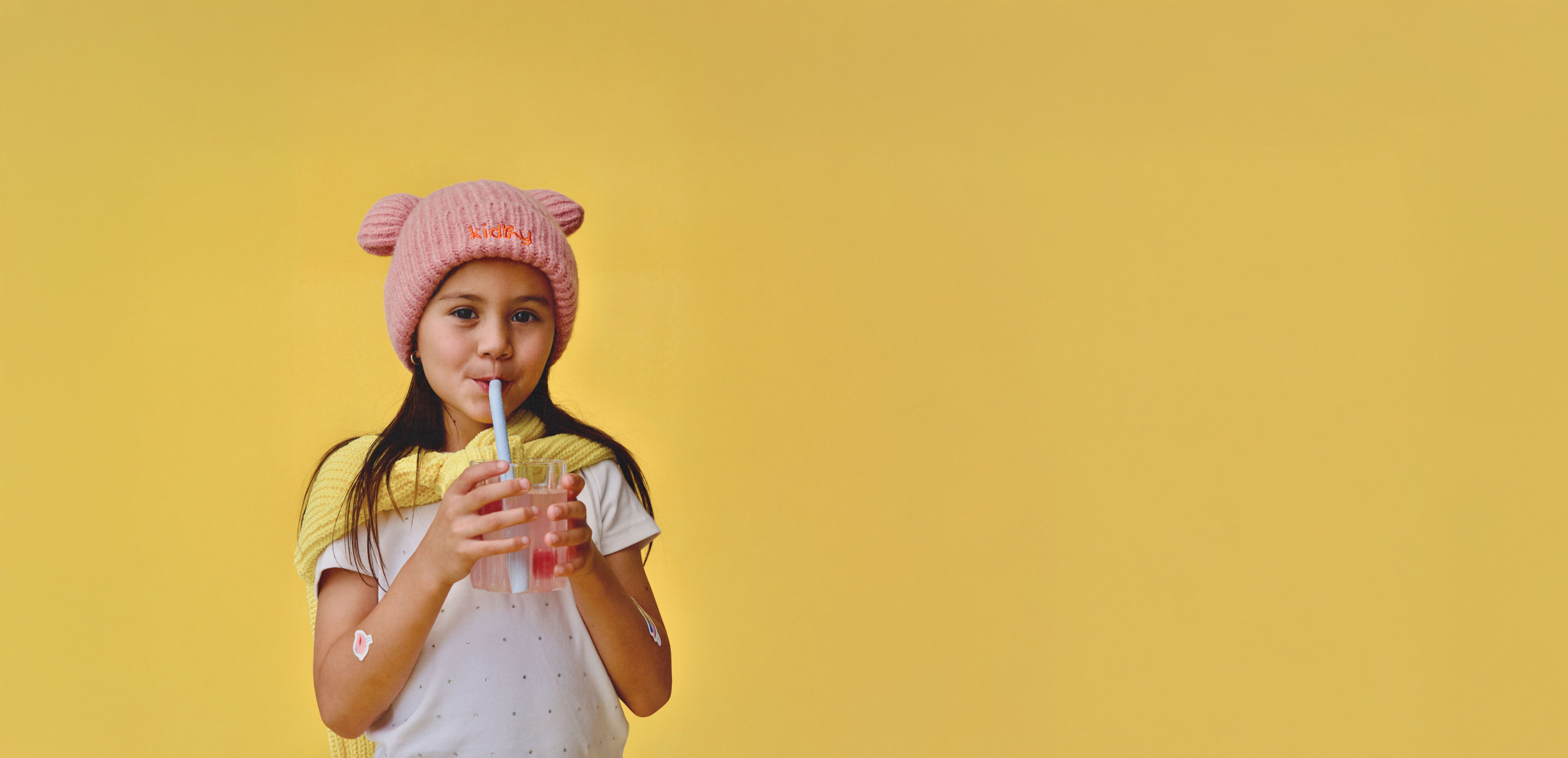 Girl with pink beanie drinking a kidhy hyration drink through a straw with a yellow background