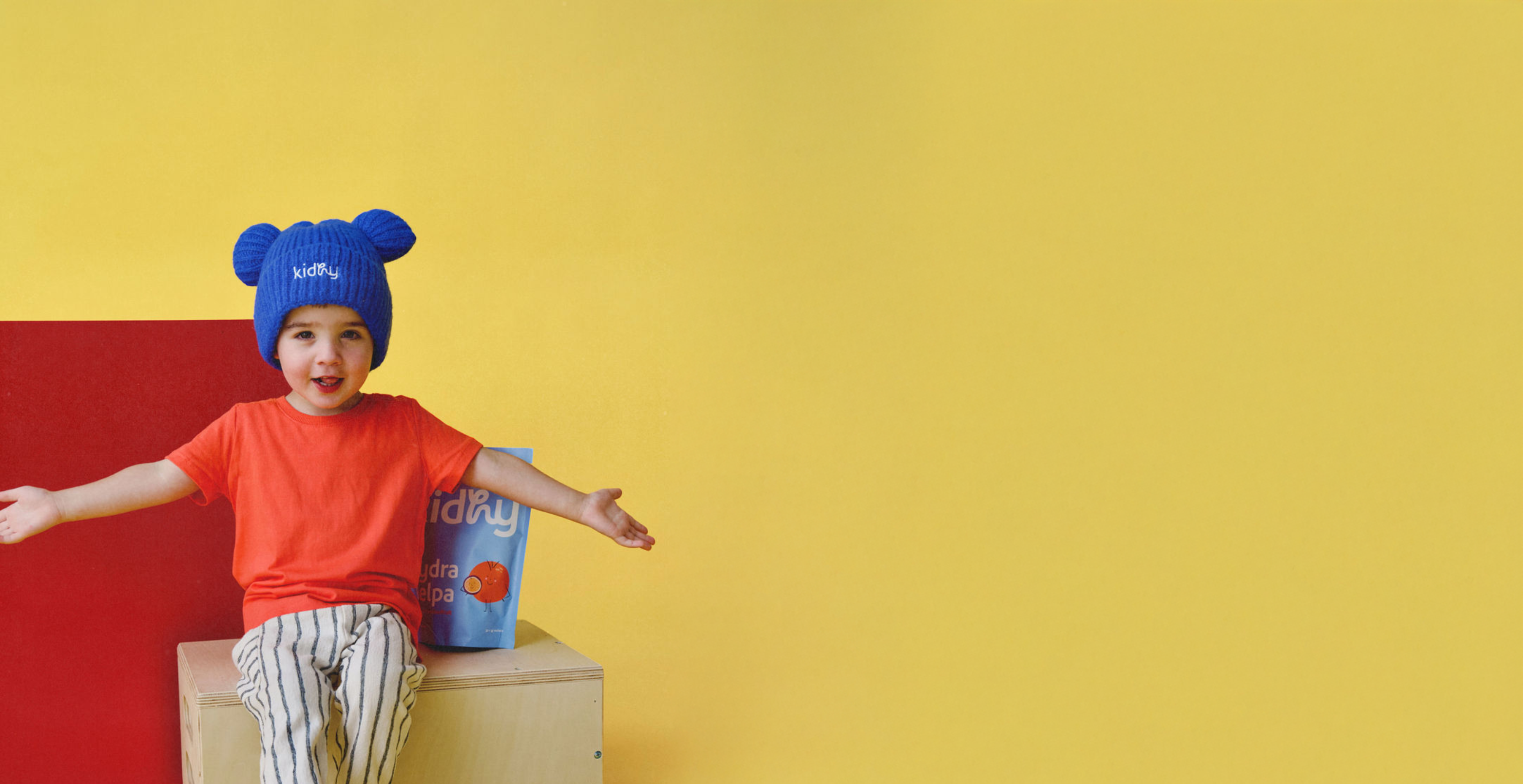 cute kid wearing a blue kidhy beanie with a pack of kidhy hyra helpa kids hydration drink in front of a yellow background