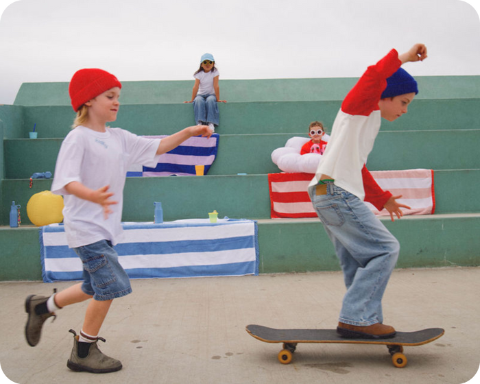 boy wearing a kidhy beanie on a skateboard being chased by another boy in a kidhy beanie, 2 kids sitting and watching them with kidhy drink bottles