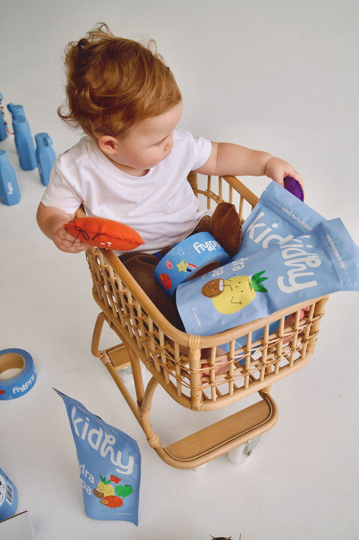 young child sitting in a toy trolley with packs of kidhy hydra helpa kids hydration drink and kidhy drink bottles in the background
